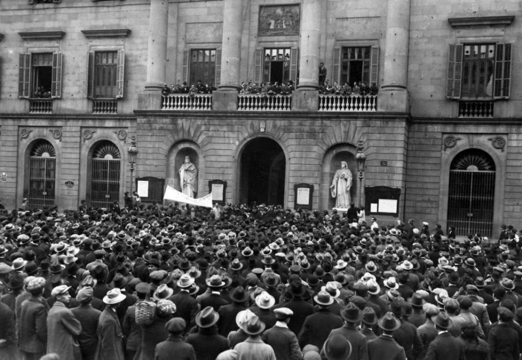 A crowd of people stand facing away from the camera looking up at a dozen people standing on the balcony of large building.