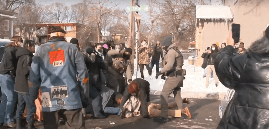 A small crowd is shown, with two ICE agents in the middle. A couple of protesters are on the ground, and one of the ICE agents appears to be swinging a baton down on the back of one of the people. A woman in the foreground has her phone out and is recording.