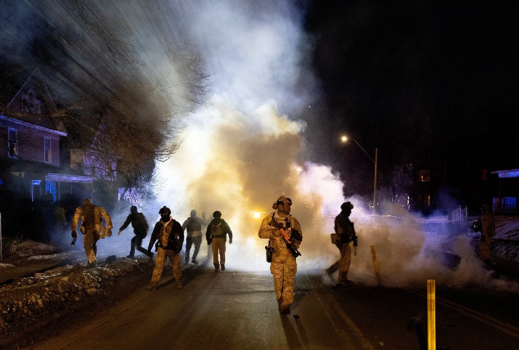 Seven federal agents in full camo and body armor walks down the street at night. A cloud of teargas behind them is illuminated by headlights.