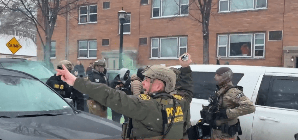 Greg Bovino with his arm cocked back holding a grenade, yelling something and about to throw it. Behind him are SUVs, other agents, and an apartment building. Green smoke is floats in the background.