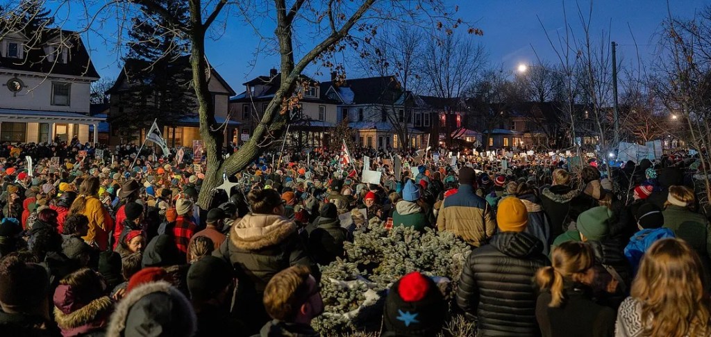 Thousands of people are standing in the street at dusk, some holding signs and others holding candles, with a line of houses in the background.
