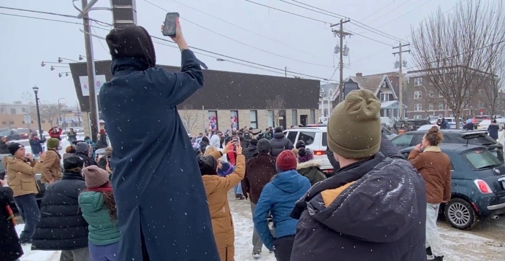 A crowd of people has their phones out and are walking after some Border Patrol SUVs. Everyone is wearing winter coats and there are flurries of snow coming down.